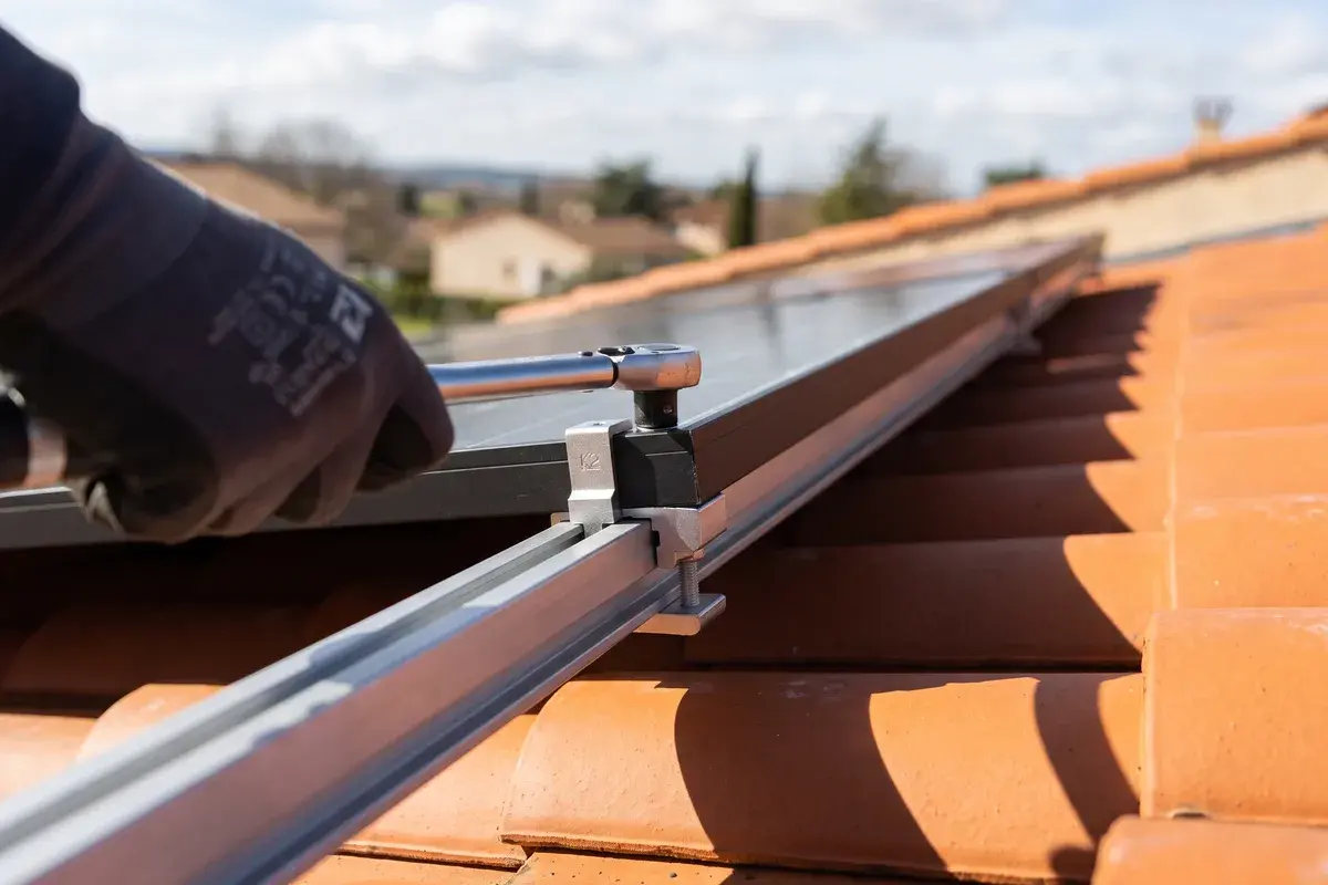 Main gantée qui serre la bride d'extrémité d'un panneau solaire sur son rail, au-dessus des tuiles