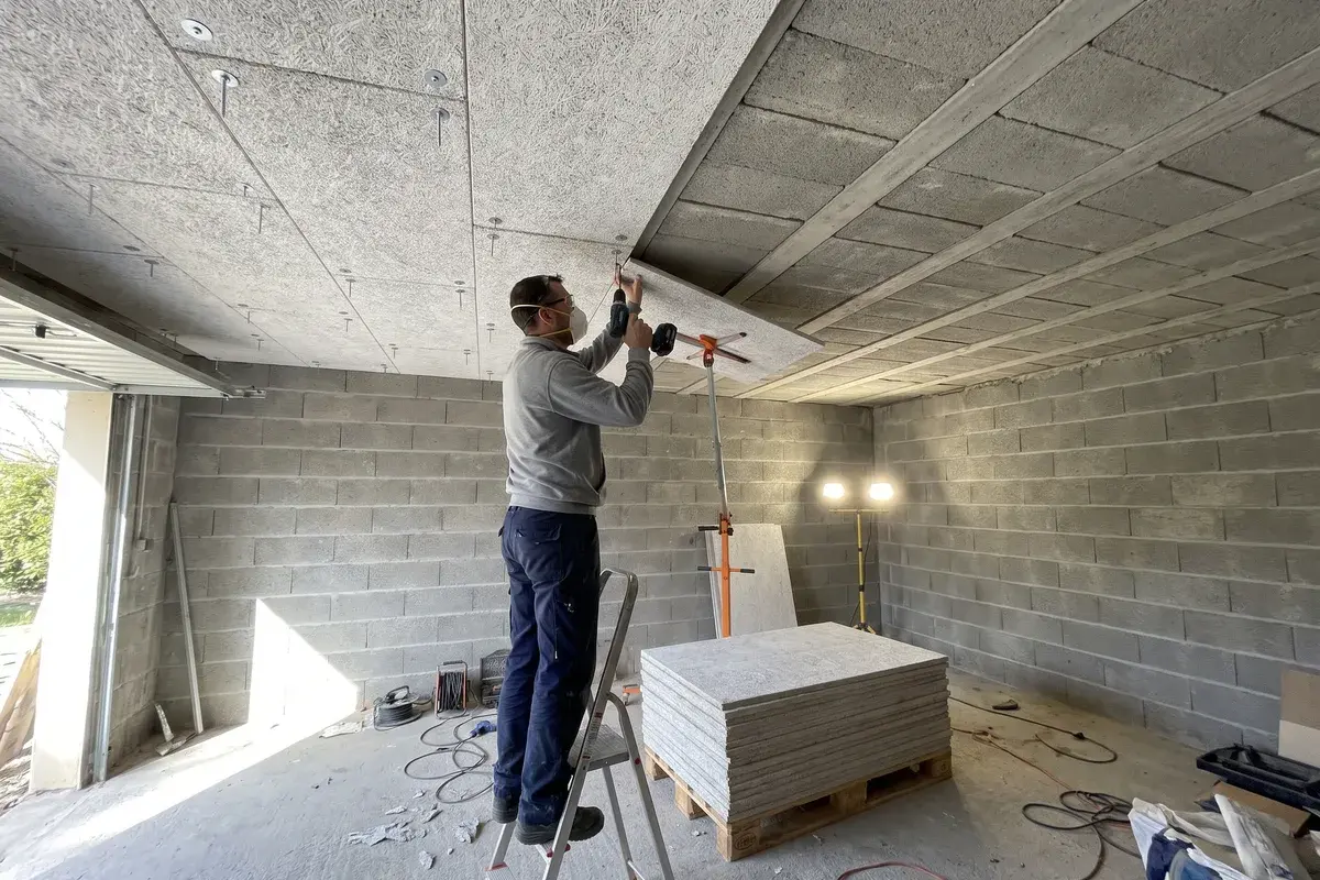 Un poseur visse des panneaux isolants Knauf Fibraroc 35 sous le plafond en poutrelles et hourdis du garage en sous-sol d’un pavillon.