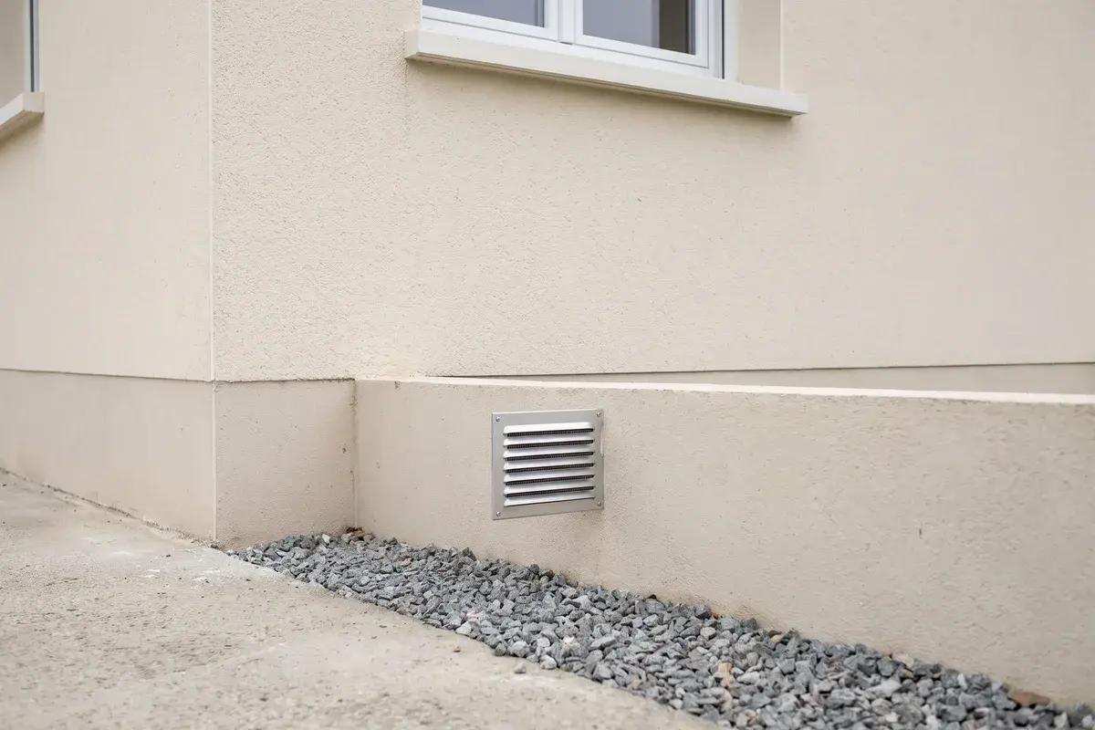 Grille d’aération métallique d’un vide sanitaire, dans le soubassement enduit d’un pavillon, au-dessus d’une bande de gravier, entièrement dégagée.