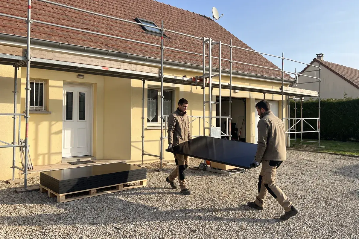 Deux installateurs portent un module monocristallin noir LONGi Hi-MO X10 vers l’échafaudage d’un pavillon en crépi jaune.