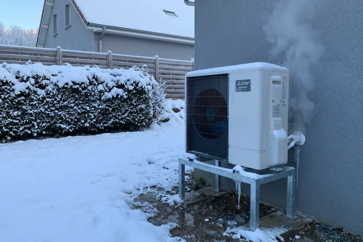 Unité extérieure Mitsubishi Ecodan sur support surélevé dans la neige, un panache de vapeur s’échappant pendant un dégivrage.