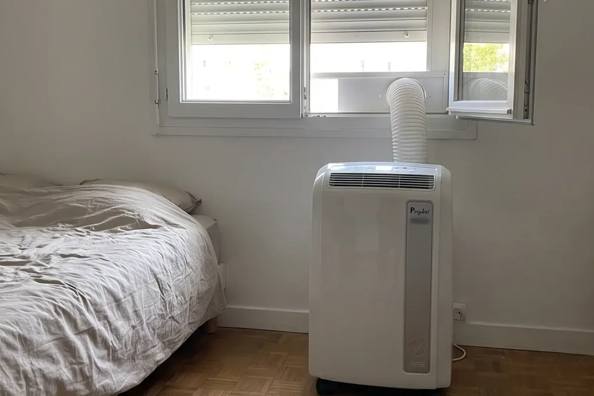 Climatiseur mobile De’Longhi Pinguino dans une chambre, sa gaine d’évacuation passée par la fenêtre entrouverte.