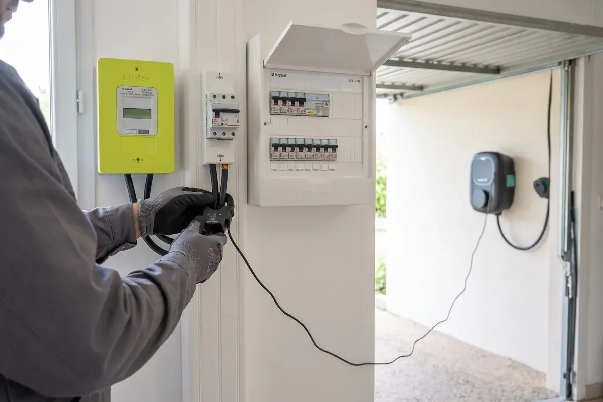 Un électricien pose une pince de mesure de courant sur le câble d’arrivée sous le compteur Linky, la borne Wallbox Pulsar Max visible dans le garage.