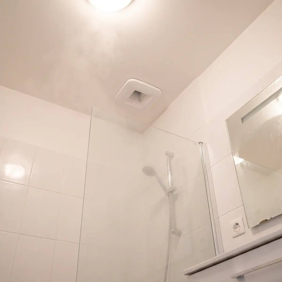 Plafond d'une salle de bains sans fenêtre : une bouche d'extraction hygroréglable Aldes Bahia Curve au-dessus de la douche, entre les murs carrelés de blanc.