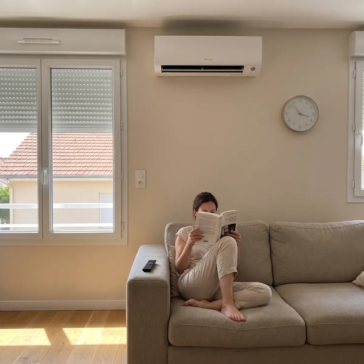 Un après-midi d'été, une habitante lit sur le canapé d'un séjour aux volets roulants à demi baissés, sous un climatiseur mural Daikin Perfera en marche.