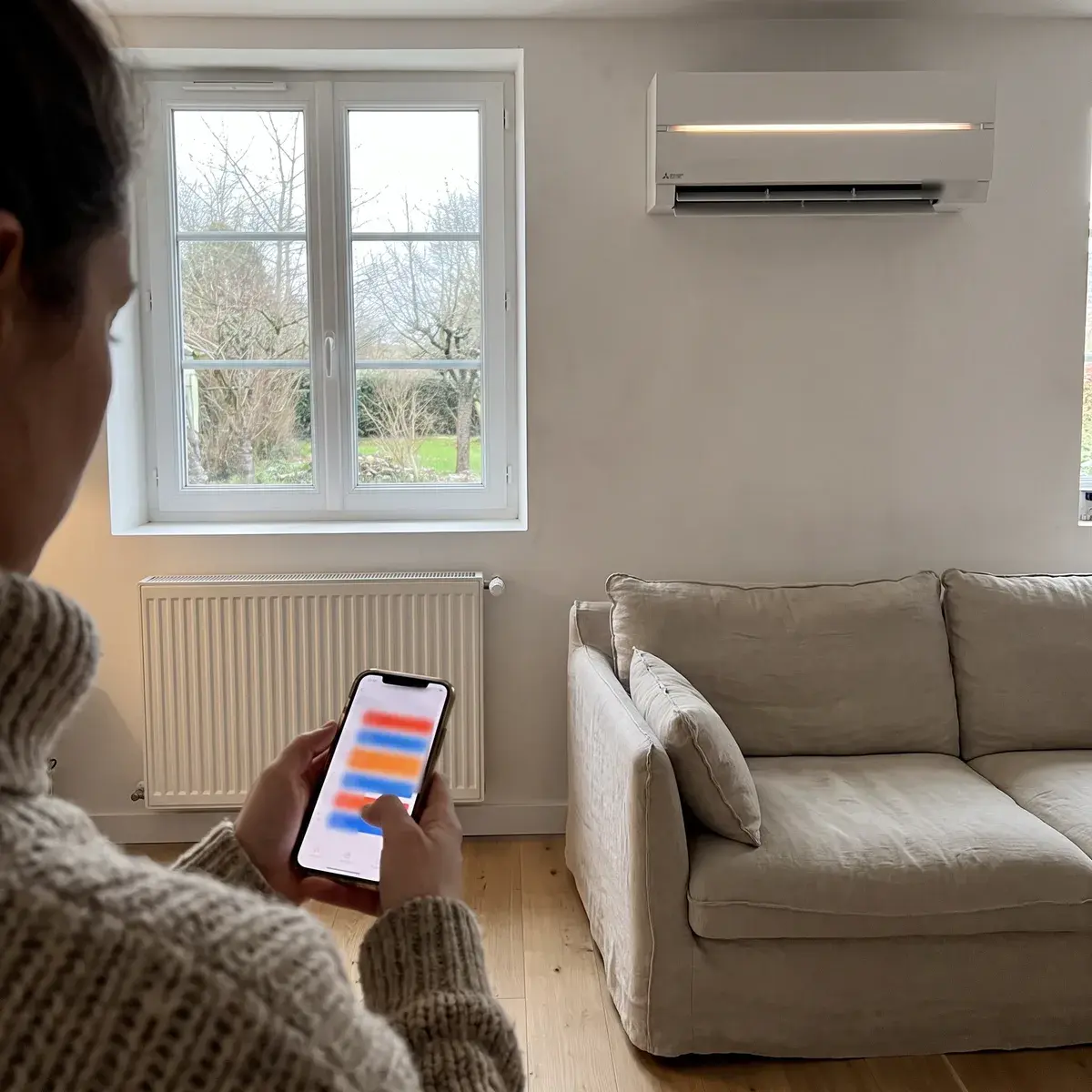 Une habitante programme des plages de chauffage sur son téléphone, sous une unité murale Mitsubishi Electric MSZ-LN, dans un séjour équipé d’un radiateur.