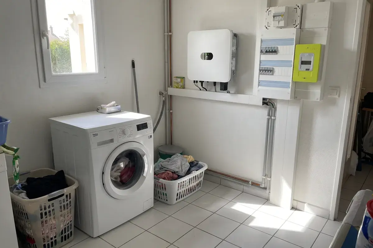 Buanderie d’une maison occupée à midi : lave-linge en marche sous le soleil, onduleur Huawei SUN2000 et compteur Linky au mur.