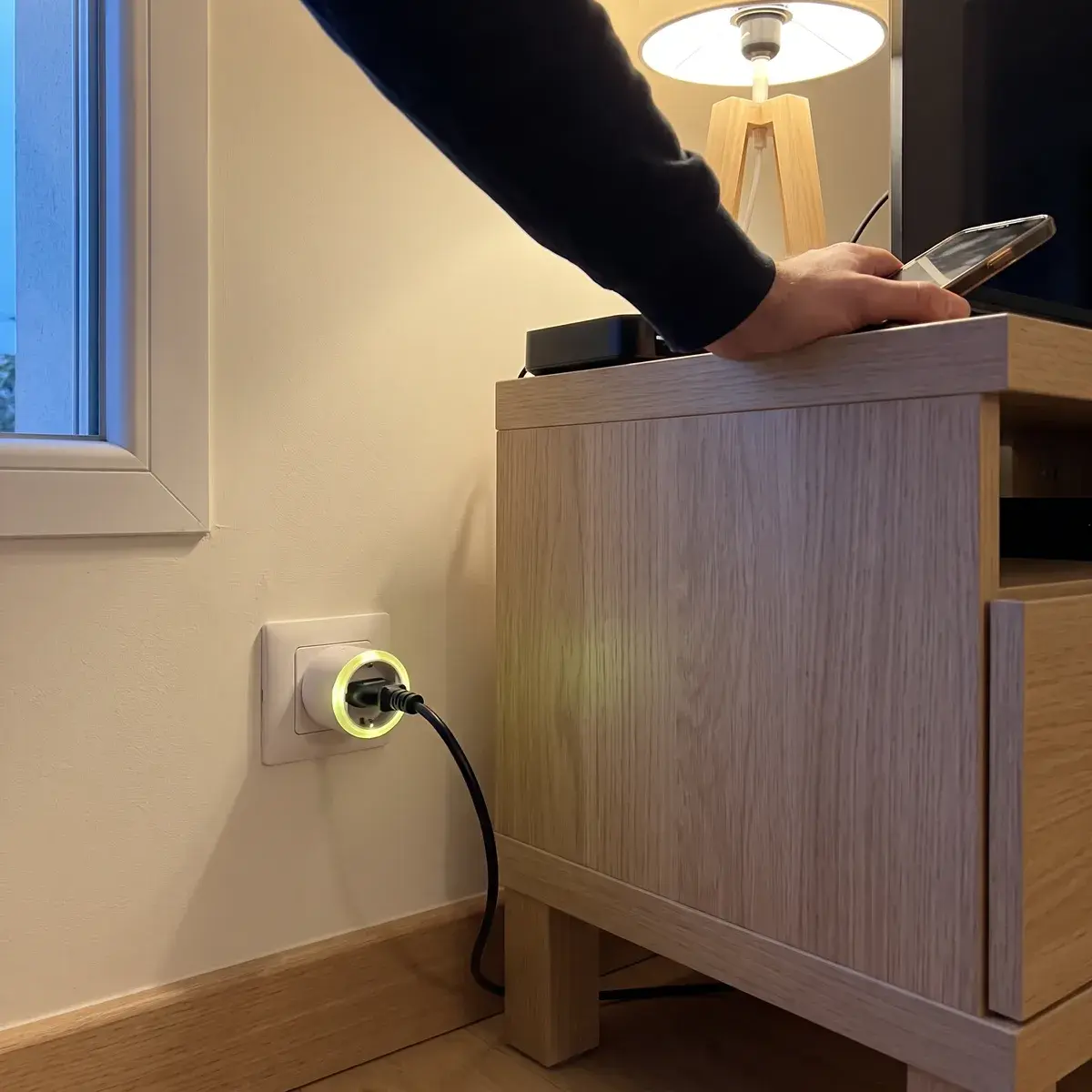 Une prise connectée Shelly Plug S, anneau lumineux vert, branchée dans une prise murale française au pied d'un meuble télé ; le câble d'un décodeur y est raccordé.