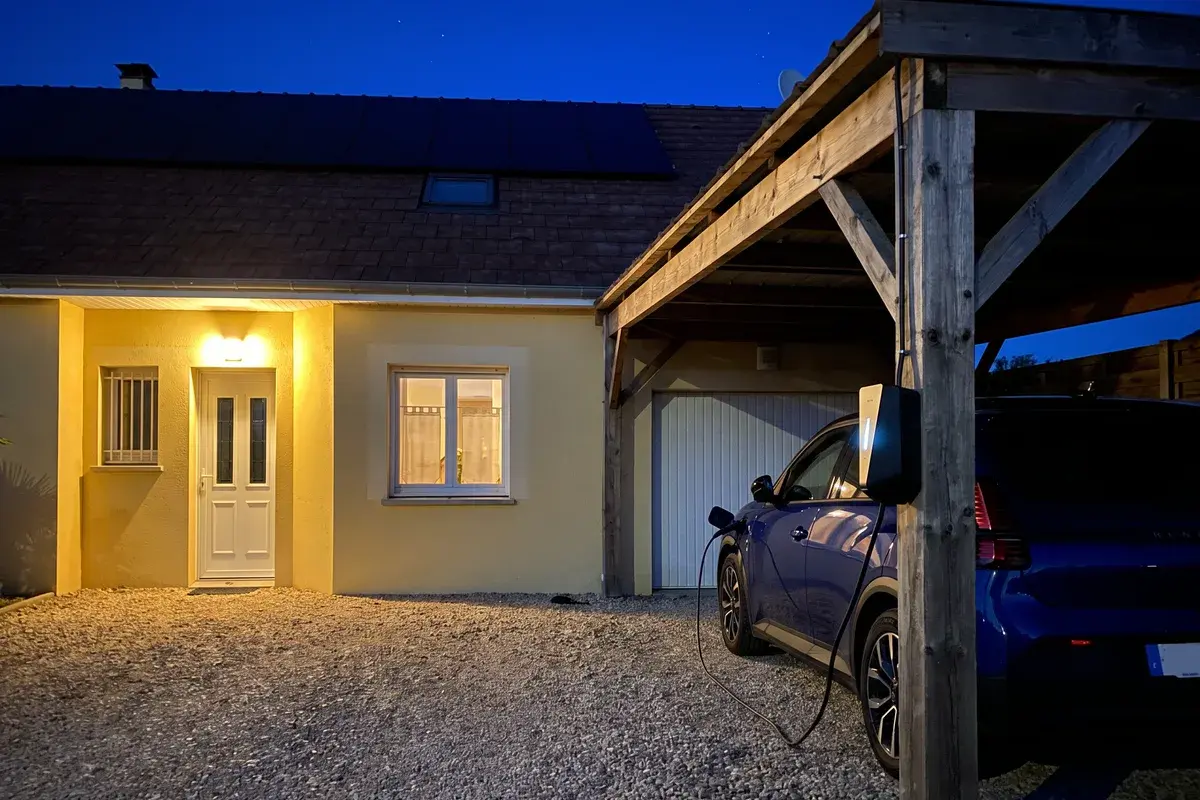 Borne Easee Charge Up fixée au poteau d’un carport en bois, la nuit, branchée sur une voiture électrique devant un pavillon en crépi.