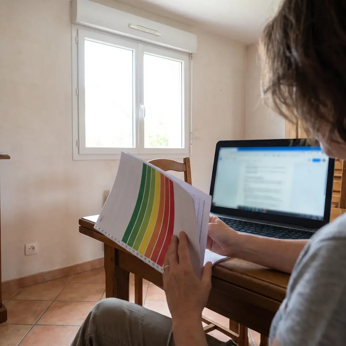Dans la salle à manger d’un pavillon, une propriétaire feuillette un rapport de diagnostic imprimé, dont la couverture montre l’échelle des étiquettes du vert au rouge, devant un ordinateur portable.