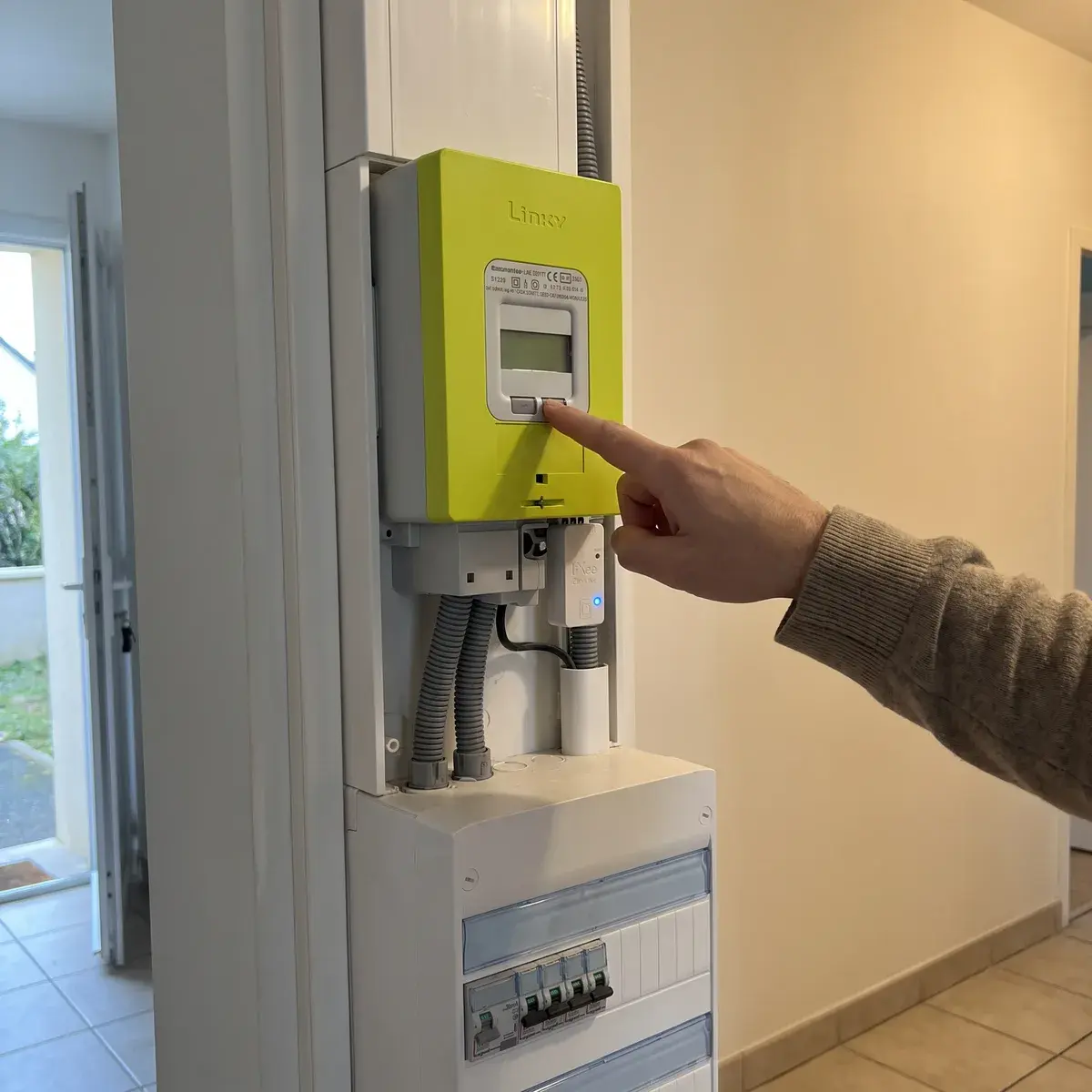 Un doigt appuie sur la touche de défilement d'un compteur Linky vert ; sous le compteur, un petit module LiXee ZLinky branché sur la sortie de télé-information, au-dessus du tableau électrique.
