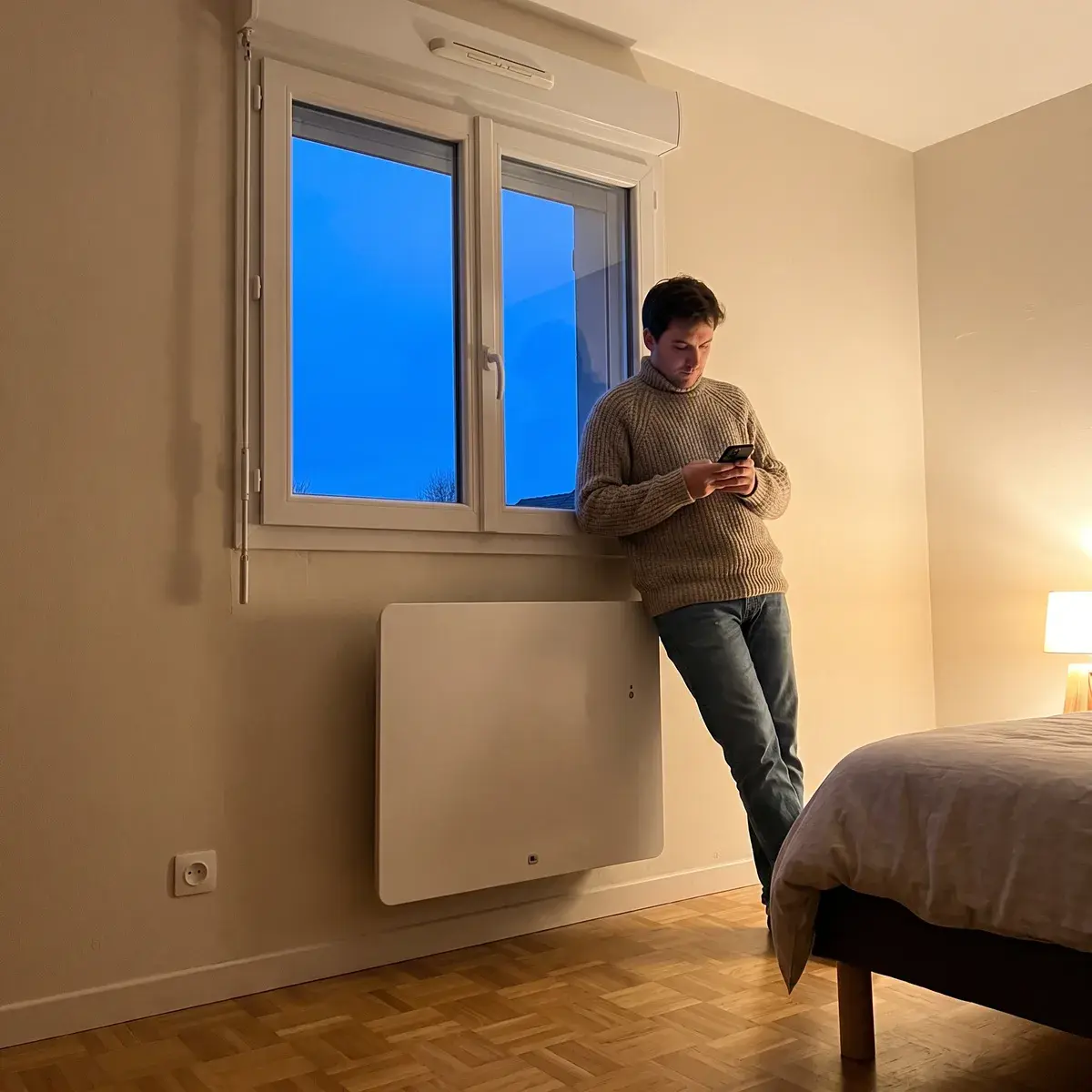 Un soir d'hiver, un homme en pull s'appuie à la fenêtre d'une chambre, au-dessus d'un radiateur électrique Thermor Équateur 5 blanc à façade lisse.