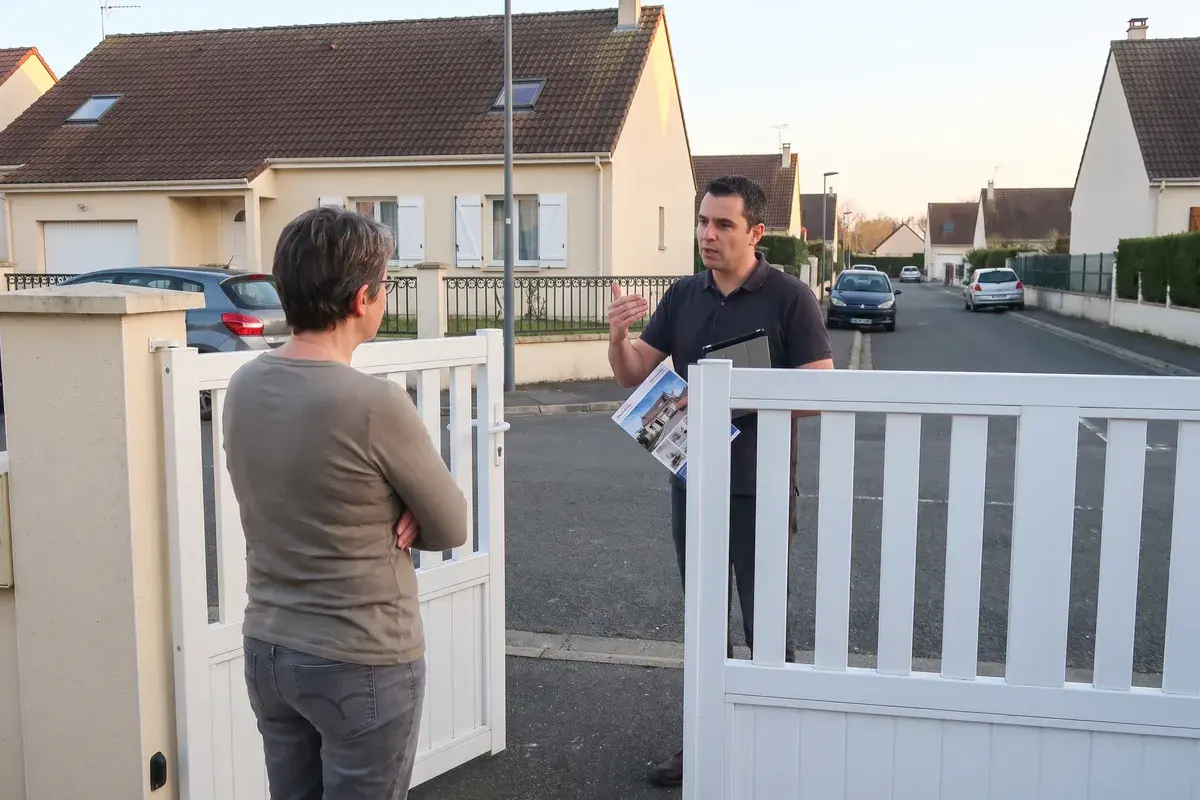 Un démarcheur muni d’une tablette et d’une brochure parle à une propriétaire restée derrière son portail, dans une rue pavillonnaire.