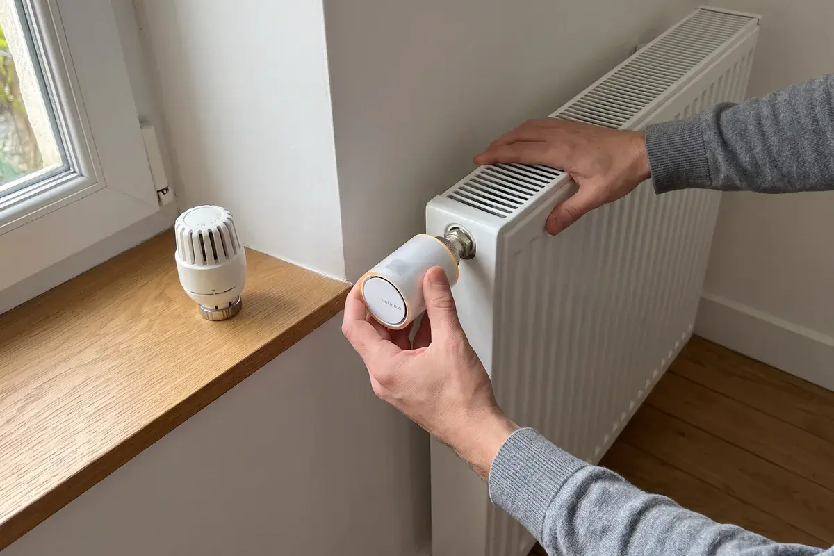 Un particulier visse une tête thermostatique connectée Netatmo sur le corps de vanne d’un radiateur en acier, l’ancienne tête manuelle posée sur l’appui de fenêtre.